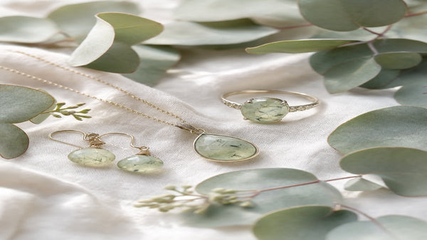 Prehnite crystal jewelry — pale sage green pendant, ring, and earrings on white linen with eucalyptus